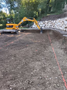 A yellow excavator operating in a dirt area near a stone retaining wall, with trees in the background and a red line marked on the ground.