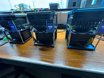 Three Bitaxe or similar open-source Bitcoin ASIC miners running on a wooden table, with digital displays showing hash rates and connected by yellow ethernet cables.