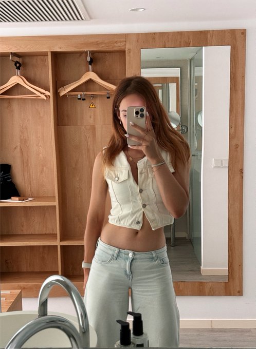 A woman takes a mirror selfie in a bathroom wearing a white sleeveless cropped vest and light blue jeans, with a wooden wardrobe visible behind her.