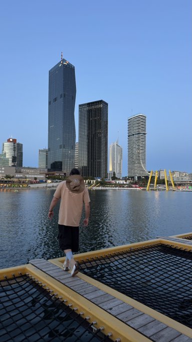 A man wearing a mask and beige shirt walks across a yellow and black floating dock on a body of water, with a city skyline of tall buildings behind him.