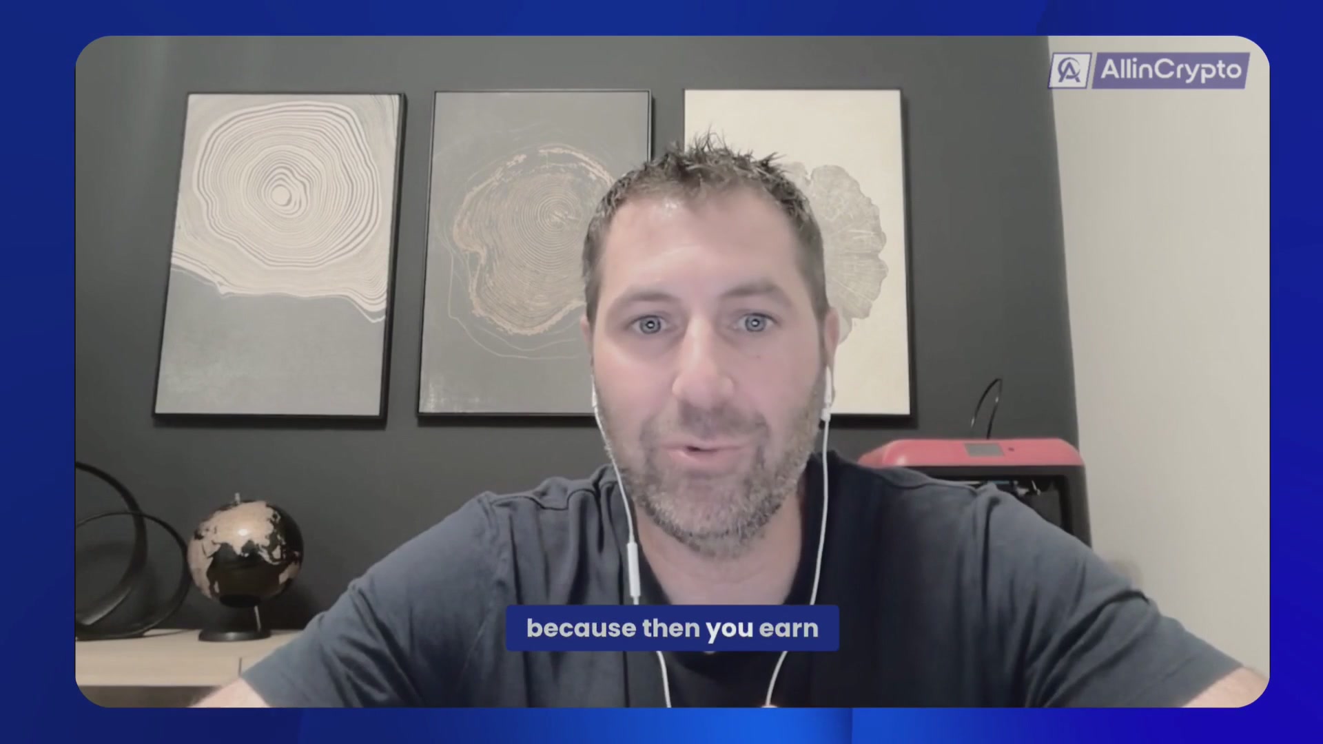 The image shows a man wearing headphones sitting at a desk with a computer screen in front of him. The man appears to be engaged in a video call, possibly discussing cryptocurrency or related topics. There are three pictures hanging above his head on the wall behind him, and a globe is also present in the room.