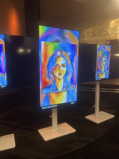 A photo from an NFT art gallery display showing multiple illuminated screens featuring a vibrant, colorful digital portrait of a woman with blonde hair, labeled "ZAZ GALLERY" and "NFT".