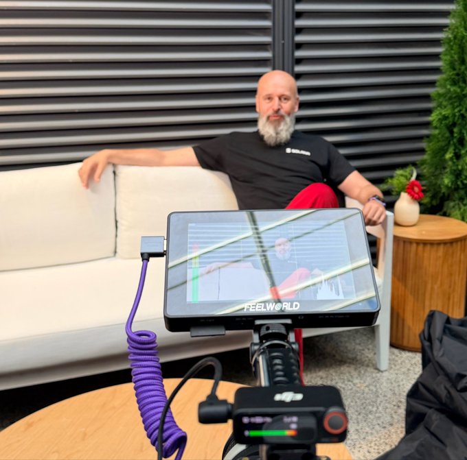 A bearded man in a black "Solana" t-shirt sits relaxed on a cream outdoor sofa while being filmed by a DJI camera with a FEELWORLD monitor displaying his live shot, with a coiled purple cable running across a wooden table in the foreground. The setup, complete with professional video equipment, humorously illustrates the speaker's claim that their elaborate efforts to motivate developers to build a Solana Mobile app have reached ridiculous extremes.