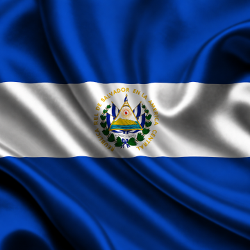 The waving flag of El Salvador features blue and white horizontal stripes with its national coat of arms in the center, encircled by the text "REPÚBLICA DE EL SALVADOR EN LA AMÉRICA CENTRAL." The image illustrates the accompanying post about El Salvador receiving a $139 million IMF tranche despite its controversial adoption of the cryptocurrency, with the country's flag symbolizing the nation at the center of this Bitcoin-IMF dispute.