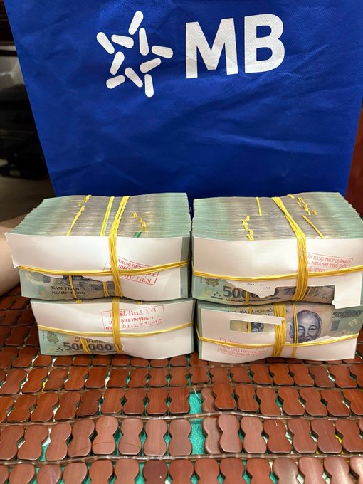 Stacks of Vietnamese 500,000 Dong banknotes bundled with yellow rubber bands, sitting on a tiled surface in front of a blue flag with 'MB' (Military Bank) logo.