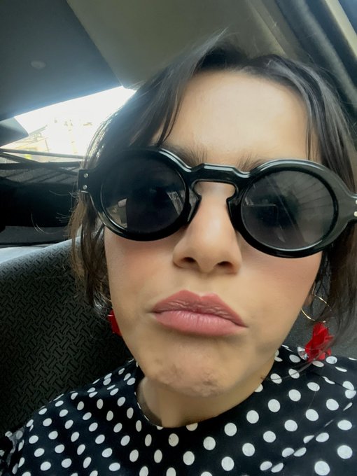 A selfie of a woman wearing oversized round black sunglasses and a black-and-white polka dot top, making a pouty expression inside a car.