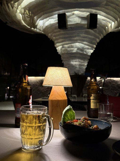 A dimly lit restaurant scene featuring a glass mug of beer, a bowl of food, two beer bottles, and a warm lamp on a table.