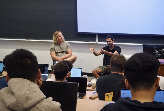 Two men sit at the front of a classroom in front of a projection screen, engaging with a group of students who have laptops open, appearing to be a tech talk or workshop session.