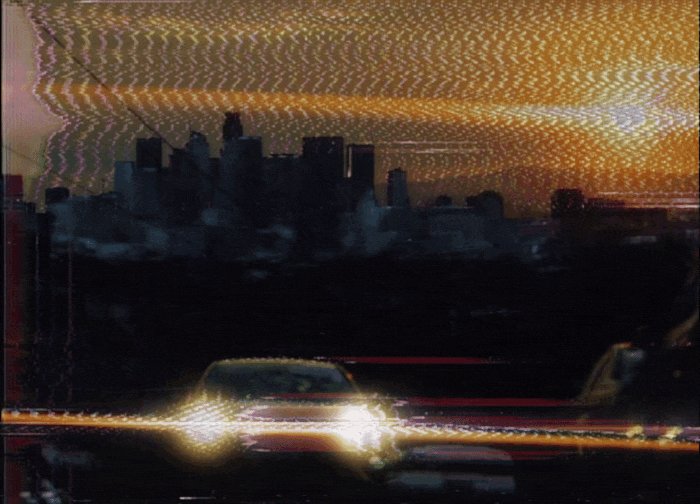 The image features an urban cityscape with a car driving down the street, surrounded by tall buildings that create a sense of depth and perspective. The sky above appears hazy or blurred due to the camera's focus, adding a dreamy quality to the scene. The photo is taken from behind the moving car, giving viewers a unique perspective on the city as it passes by.