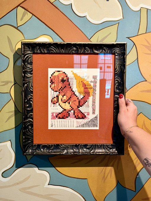 A framed pixel art piece depicting a dinosaur, being held against a floral-patterned wall by a person with red nail polish and tattoos.