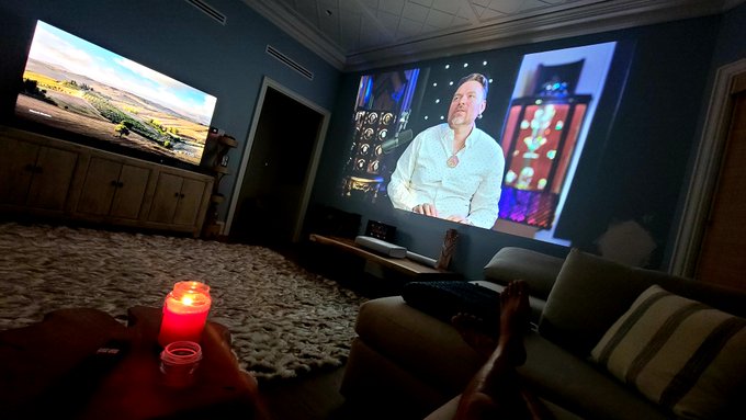 A cozy living room features a large projection of a man in a white shirt, with a TV displaying a scenic landscape and a lit candle on a coffee table in the foreground, creating an intimate viewing atmosphere for what appears to be a story about Richard Heart. The projection screen suggests viewers are watching a documentary or presentation about him, visually reinforcing the post's narrative of people judging him before knowing his full story.