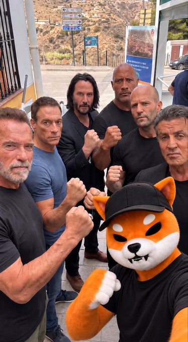 An AI-generated group photo shows several muscular men, including likenesses of Sylvester Stallone, Dwayne Johnson, and Jason Statham, posing with clenched fists alongside an anthropomorphic orange Shiba Inu mascot wearing a black cap, standing on a sunny street.