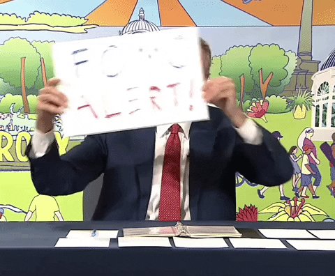 The image features a man wearing a suit, holding up a sign that reads "FOF TALERT!". The man appears to be sitting at a table with papers or documents spread out before him. The background of the image includes an illustration depicting a city scene and a person riding a bicycle.