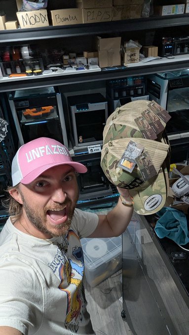 A selfie of an excited man in a pink 'UNFAZED' cap holding up a camouflage 'Trucker' cap in what appears to be a 3D printing workshop.