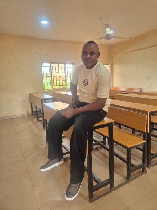 A man sits on a school desk in a classroom, wearing a t-shirt with "WE ARE" text, smiling at the camera in what appears to be an educational setting. The classroom environment with empty desks and benches contrasts with the post's discussion of privacy-focused programmable technology like Beldex's Confidential EVM research.