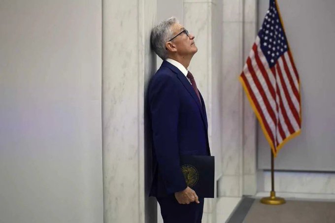 Federal Reserve Chair Jerome Powell, wearing a blue suit and glasses, leaning against a marble wall and looking upward while holding a folder, with an American flag in the background.
