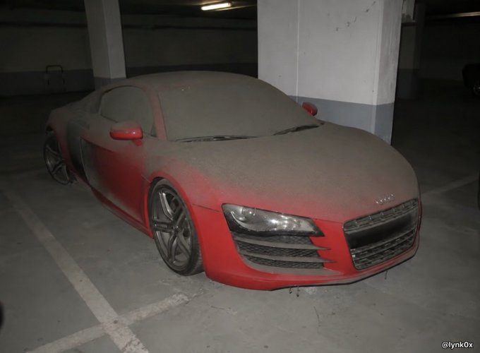 A red Audi sports coupe covered in thick dust sits parked inside a dim concrete garage.