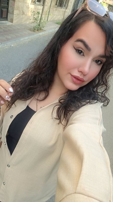 A woman with long curly dark hair and sunglasses on her head takes a selfie on a street, wearing a cream-colored cardigan over a black top with a necklace.