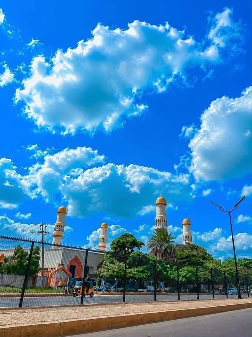 A bright blue sky filled with fluffy white clouds over a mosque with multiple minarets and palm trees, viewed from behind a chain-link fence.