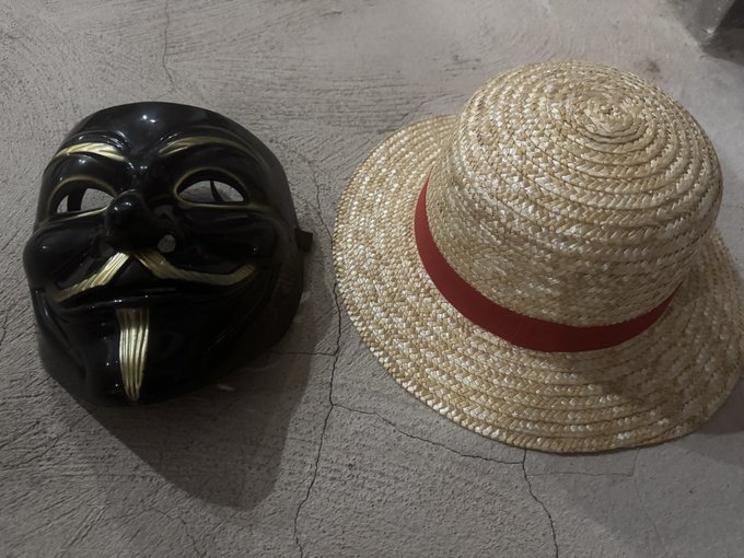 A black mask with gold facial detailing sits beside a straw fedora with a red band, embodying the post's message of taking on new personas and pursuing bold challenges—just like Luffy from One Piece, the mask resembles his iconic grin, symbolizing perseverance and never giving up.