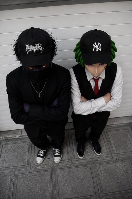 Two people pose back-to-back; one wears a black cap with death metal band logo and a mask, while the other sports a green wig, a NY Yankees cap, and a formal vest with red tie.