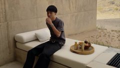 A man in a beanie, grey shirt, and dark pants sits on a concrete bench in a modern room, resting his chin on his hand next to a small table with fruits.