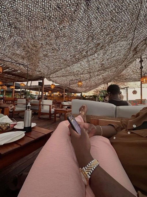 A first-person perspective from a lounge seat under a woven canopy roof, showing the photographer's legs, hand holding a phone, and a gold watch, with other patrons relaxing on couches in a tropical restaurant setting.