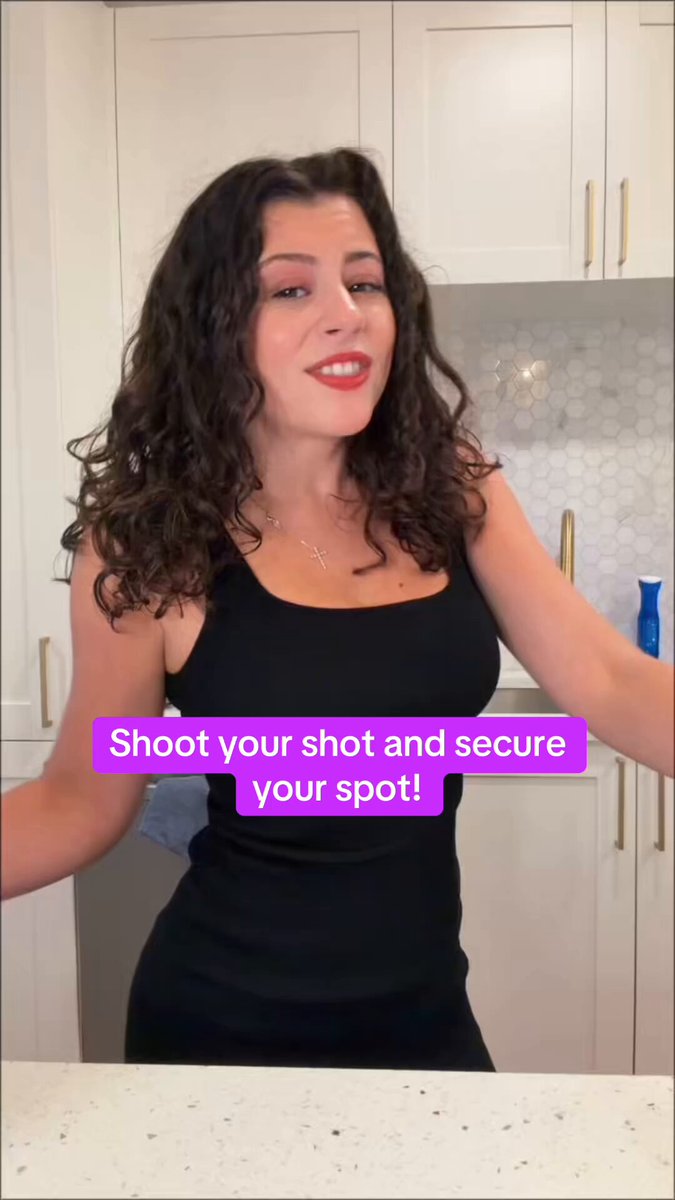 The image features a woman standing next to a kitchen counter, wearing a black dress. She has curly hair and is smiling for the camera. The background of the photo shows white cabinets and a sink, indicating that she might be in her own home or a kitchen setting. There are also some text elements in the image, including "SHOOT YOUR SHOOT AT A FUTURE WITH BIP44", which could possibly be related to Bitcoin or cryptocurrency.