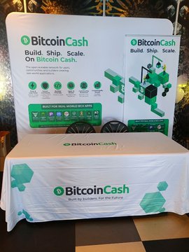 A Bitcoin Cash promotional booth setup with branded banners reading 'Bitcoin Cash - Build. Ship. Scale.' and a table displaying the same branding, shown at what appears to be a convention.