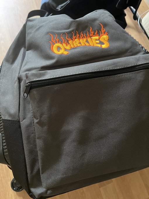 A black bag featuring "QUIRKIES" embroidered in flame letters sits on a wooden floor, which the poster is showing off as their "Quirkies NFT backpack" before heading to the grocery store.