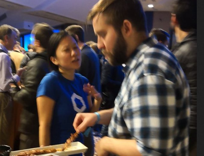A photograph from what appears to be an indoor event showing a bearded man in a blue and white plaid shirt holding a tray of food (skewers), with a woman in a blue Digit logo shirt standing next to him, surrounded by other people.