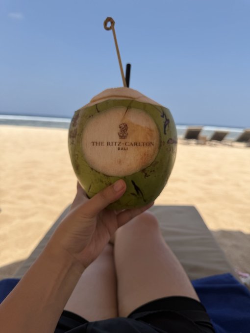 A person's hand holding a fresh green coconut with a straw, branded 'THE RITZ-CARLTON BALI', while relaxing on a beach lounger looking out at the ocean.