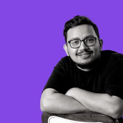 A black-and-white portrait photo of a smiling man with glasses and a beard, arms crossed, set against a purple background.