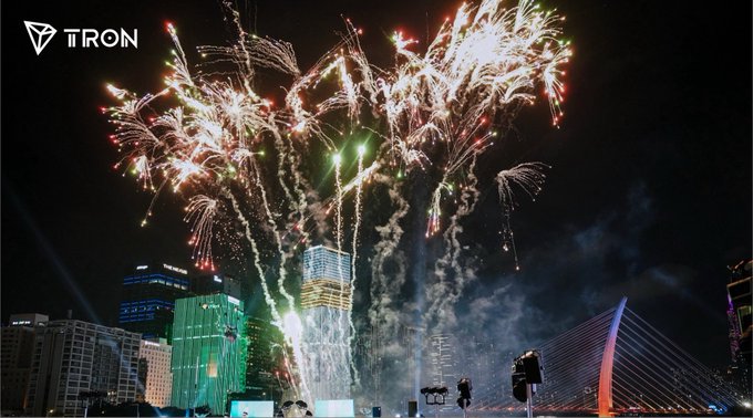 A nighttime photograph of a large, colorful fireworks display exploding over a city skyline, featuring the 'TRON' logo in the top left corner.