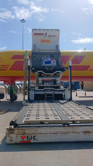 A DHL cargo plane is being unloaded at an airport, with workers using a conveyor belt loader to transfer packages, including pallets labeled "MEDIA +" and "DHL," symbolizing the arrival of aid donations from Panama to Venezuela. The image visually reinforces the message that humanitarian donations have successfully reached their intended destination.