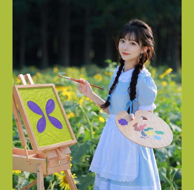 A young woman in a light blue dress stands in a sunflower field holding a paint palette and brush, smiling beside an easel displaying a small painting of a purple flower.