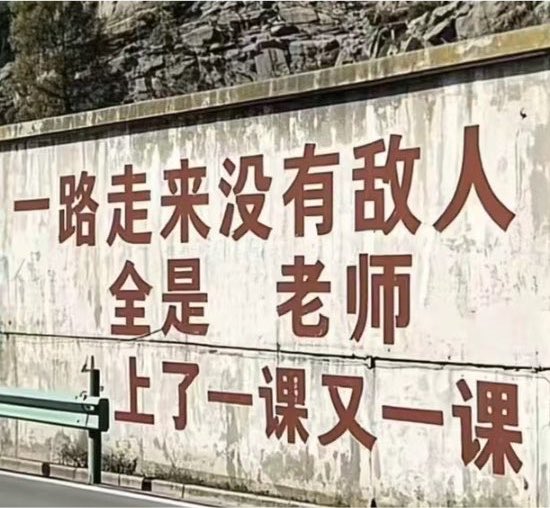 Red Chinese characters are painted on a concrete wall along a road, reading '一路走来没有敌人 全是老师 上了一课又一课' (Throughout the journey there were no enemies, only teachers, lesson after lesson).