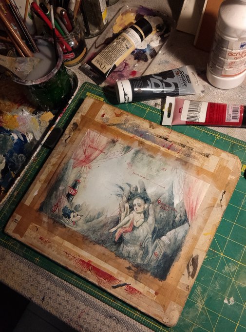 An artist's workspace showing a watercolor or mixed media painting in progress featuring a figure with flowing hair, surrounded by tubes of acrylic paint, brushes, and a green cutting mat.