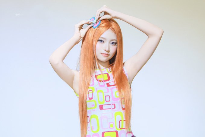 A young woman with long, straight orange hair poses with her hands raised above her head, adjusting a small blue and orange hair accessory. She wears a sleeveless dress featuring a colorful geometric pattern in shades of pink, yellow, and green against a soft, gradient pastel background. A small watermark reading "AG" is visible in the upper left corner.