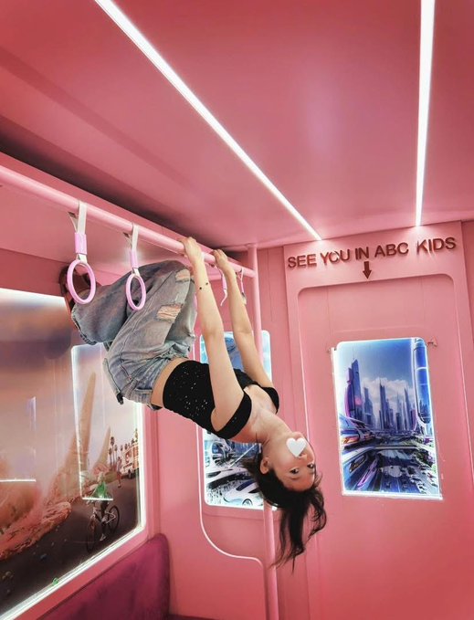 A young woman hangs upside down from gymnastic rings inside a pink train or subway car decorated with the phrase 'SEE YOU IN ABC KIDS' and futuristic cityscape posters.