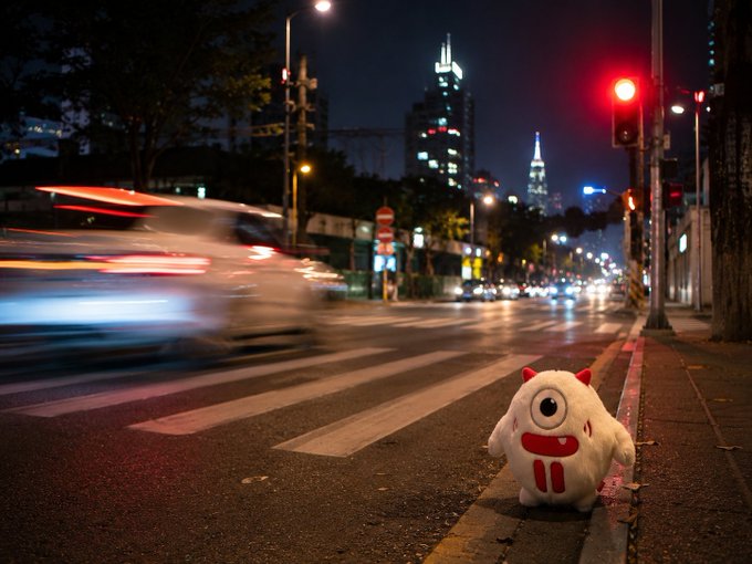 A plush toy sits on a city street at night with blurred car lights streaking past and a red traffic signal illuminating the scene.