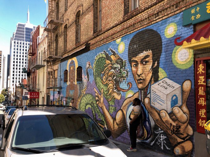 A vibrant Bruce Lee mural in an urban Chinatown setting features the martial artist holding a dragon and a box labeled 'Chain Link' with a padlock, with Chinese characters and McDonald's arches visible on the brick wall while a person and cars occupy the street below.