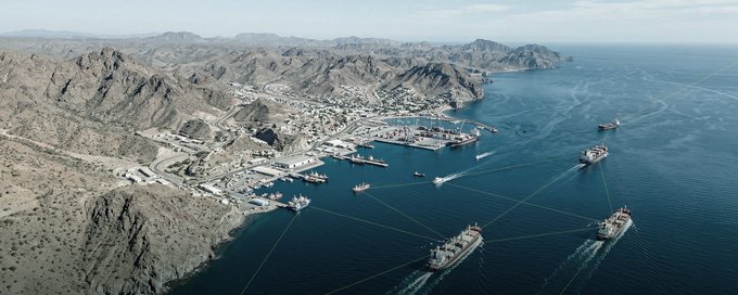 Aerial view of a coastal harbor with ships and port infrastructure nestled between arid mountains and the ocean, overlaid with a digital network graphic showing connected nodes.