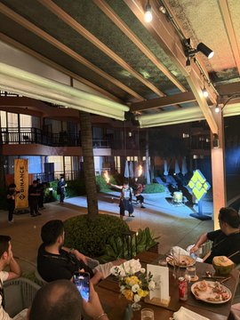 An evening outdoor scene at what appears to be a beachside restaurant or bar, with people seated at tables in the foreground while others walk past closed beach umbrellas and a yellow flag in the background.