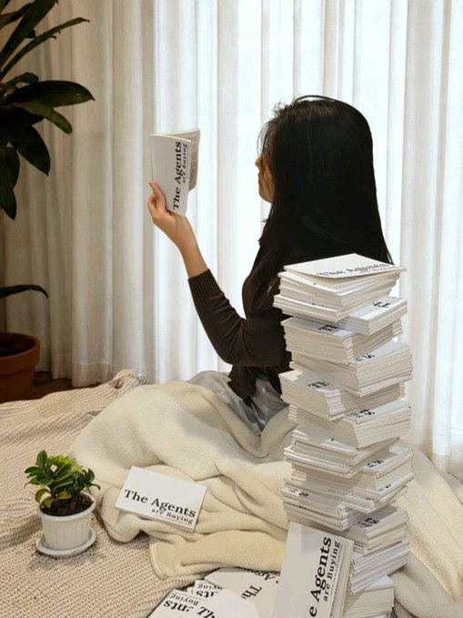 A woman sits on a bed reading "The Agents Are Buying" while surrounded by multiple stacks of the same book, visually emphasizing the excitement of unveiling her new pocketbook titled "The Agents Are Buying: Field Notes from the Onchain Agentic Economy 2026" at Token2049 Singapore.