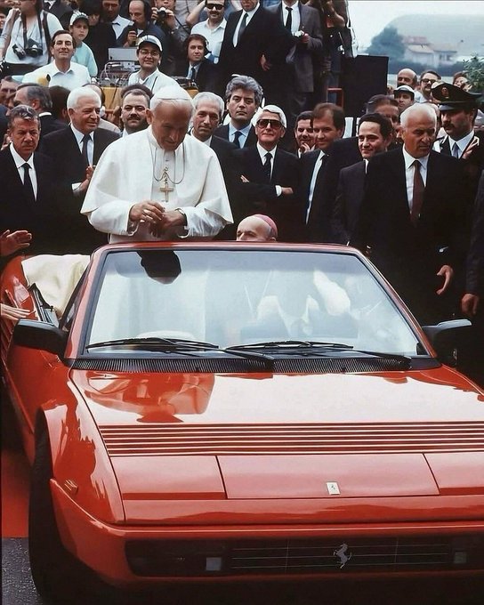 Pope John Paul II, dressed in white vestments, stands in a red Ferrari convertible surrounded by a crowd of officials, bodyguards, and onlookers.