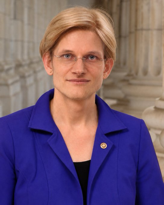 A woman with short blonde hair, wearing glasses and a bright blue blazer over a black top, stands in front of what appears to be a classical stone-columned building interior. A small gold pin is visible on her lapel. The image appears to be an official portrait, consistent with the post referencing Elizabeth Warren, the U.S. Senator known for her signature blue attire.