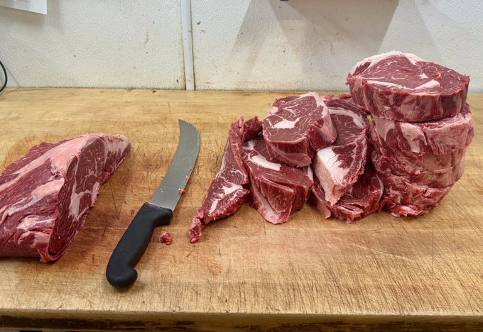 Raw ribeye steaks sliced into thick portions on a wooden butcher block cutting board next to a black-handled boning knife.