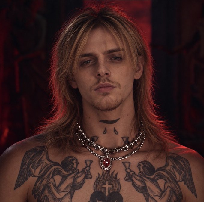 A shirtless young man with long blonde hair, a nose ring, and extensive tattoos including angels, a cross, and a crown of thorns wears a silver chain necklace against a red-lit background.