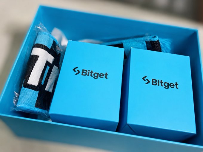 Two Bitget-branded light blue boxes sit inside a matching tray, accompanied by a rolled-up towel or wristband featuring the Bitget logo, all in the brand's signature blue and black colors. The merchandise aligns with the post expressing appreciation for a thoughtful Bitget gift, showcasing the promotional items received from the crypto exchange.